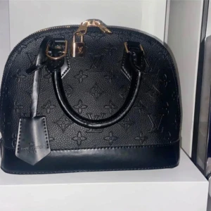 Svart handväska från Louis Vuitton - Säljer en svart alma  från Louis Vuitton. Väskan har guldfärgade detaljer och en praktisk dragkedja. Insidan är klädd i rött tyg. Pris kan diskuteras.💗