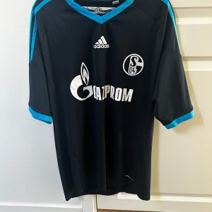 Svart fotbollströja från Adidas - Säljer en snygg svart fotbollströja från Adidas med Schalke 04-logga och Gazprom-tryck på framsidan. Tröjan har korta ärmar och turkosa detaljer vid krage och ärmslut. Perfekt för fans av Schalke 04 eller för den som vill ha en stilren sporttröja. Passar bra till både träning och vardag.