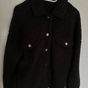 Svart teddyjacka - Mysig svart teddyjacka med knappar framtill och två bröstfickor. Perfekt för kyliga dagar när du vill vara både varm och stilren. Jackan har en klassisk krage och långa ärmar. Passar bra till både vardag och fest!