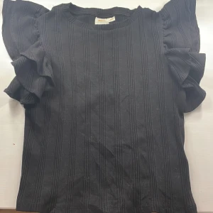 Svart ribbad topp från Gina Tricot - Säljer en snygg svart ribbad topp från Gina Tricot. Den har korta ärmar med volangdetaljer som ger en extra touch. Perfekt för både vardag och fest! 🖤