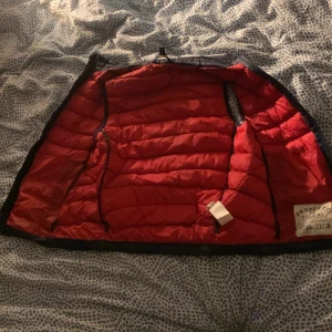 Svart dunväst från Polo RL - Säljer en snygg svart dunväst från Polo RL med röd insida. Västen har en dragkedja framtill och två praktiska fickor med dragkedjor. Perfekt för höst och vår när du vill hålla värmen utan att bli för varm. Den har en klassisk stil med det ikoniska Polo-loggan broderad på bröstet.