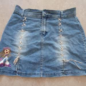 Säljer en supercool jeanskjol från Bratz med ett snyggt snörningsdetalj framtill och en färgglad Bratz-figur på sidan. Kjolen har en klassisk blå jeansfärg och två bakfickor. Perfekt för en avslappnad och trendig look!