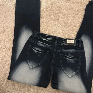 unika långmidjade jeans  - Säljer ett par mörkblå jeans med coola blekta detaljer. De är bootcut och lågmidjadd. Jeansen har fickor både fram och bak samt en klassisk dragkedja och snygga detaljer som knapparna och det blekta! midjemått:36cm innersben: 79/80cm