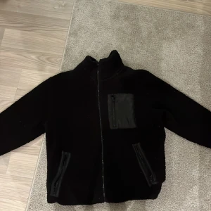 Svart teddyjacka med dragkedja - Mysig svart teddyjacka med dragkedja fram och en praktisk bröstficka. Perfekt för kyliga dagar när du vill hålla dig varm och bekväm. Jackan har en hög krage och långa ärmar, vilket gör den idealisk för höst och vinter. Den är i nyskick då jag bara använt den Max 3 ggr! Pris kan diskuteras vid snabb affär!