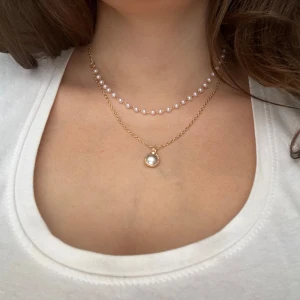 Dubbelhalsband med pärlor  - Snyggt dubbelhalsband med en kedja i guldton och en rad vita pärlor. Det ena halsbandet har en enkel pärla som hänge, vilket ger en elegant touch. Perfekt för både vardag och fest!