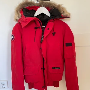 Röd dunjacka från Canada Goose - Säljer en snygg röd dunjacka från Canada Goose i mycket bra skick. Jackan har en fluffig pälsfodrad huva och praktiska fickor. Perfekt för kalla vinterdagar! 🧥❄️ Ärmmuddarna har blivit slitna och jackan är något smutsig. Storlek 2XS, skulle säga att den passar dam XS.
