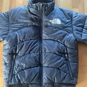 Svart pufferjacka från The North Face - Säljer en snygg och varm svart pufferjacka från The North Face. Jackan har en dragkedja framtill och två praktiska fickor med dragkedjor. Perfekt för kalla vinterdagar och höstpromenader. Den är i bra skick 8/10 men en liten skada på dragskedjan.