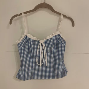 Blå och vit rutig linnetopp - Söt blå och vit rutig linnetopp med smala axelband och volangdetaljer vid halsringningen. Toppen har en knytning framtill och är perfekt för middagar eller fester. 