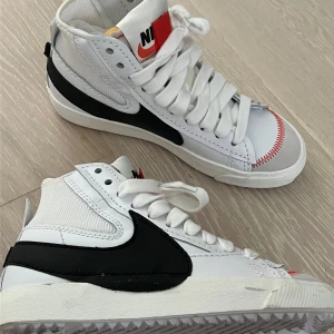 Nike 38 - Säljer ett par Nike Blazer Mid '77 i vitt och svart. De har en cool retrostil med hög snörning och en röd detalj vid tån. Perfektför vardag 🖤