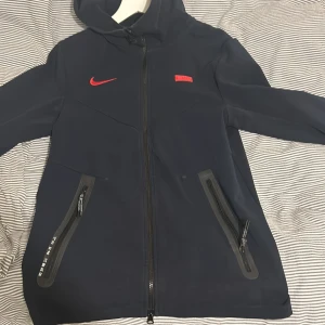 Nike tech fleece Frankrike jacka  - Säljer en mörkblå jacka med huva från Nike tech fleece. Den har röda detaljer som loggan och en emblem på bröstet. Jackan har dragkedja och två fickor med dragkedjor. Perfekt för höst och vår, och den är i mycket bra skick. Passar både till vardags och sportiga aktiviteter.