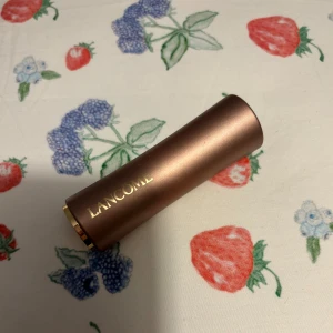 Läppstift från Lancôme - Säljer ett elegant läppstift från Lancôme i en lyxig roséguldig hylsa. Färgen är en vacker, dämpad rosa som passar perfekt för både vardag och fest. Läppstiftet har en krämig konsistens som återfuktar läpparna och ger en jämn applicering. Perfekt för att lägga till en touch av glamour till din look!