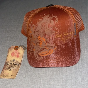 Ed Hardy keps - Säljer en jätte fin vintage orange keps från Ed Hardy. Kepsen har en justerbar baksida och en snygg dödskalle-detalj i metall. Helt oanvänd. Originellt pris var 500kr. Pris kan diskuteras.