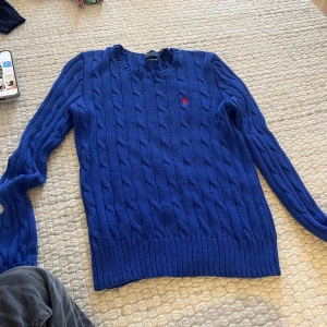 Blå stickad tröja från Ralph Lauren - Säljer en snygg blå stickad tröja från Ralph Lauren i pima bomull. Den har ett klassiskt flätmönster och en liten röd logga på bröstet. Perfekt för både höst och vinter, med långa ärmar och ribbade kanter. Superbekväm och stilren! 💙