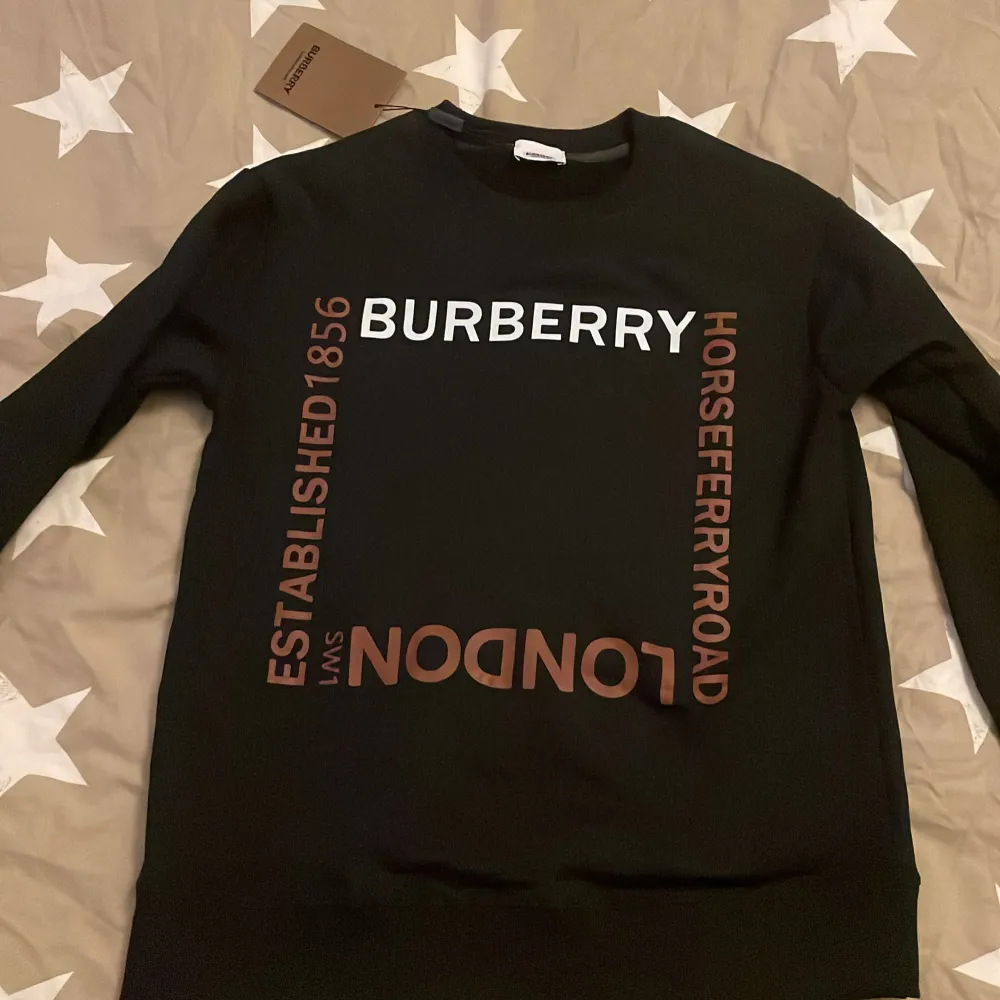 Säljer en snygg svart tröja från Burberry med tryck i vitt och brunt. Tröjan har en klassisk rund hals och är perfekt för både vardag och fest. Den är i nytt skick och har en stilren design med texten 'Burberry' och 'London' tryckt på framsidan.. Tröjor & Koftor.