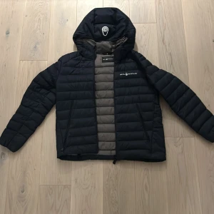 Blå pufferjacka från Sail Racing - Säljer en svart pufferjacka från Sail Racing i mycket bra skick. Jackan har en quiltad design med dragkedja och är perfekt för höst och vinter. Den är långärmad och har en snygg logga på bröstet. Perfekt för att hålla sig varm och stilren! Den har inte någon defekt storlek 160cm barn