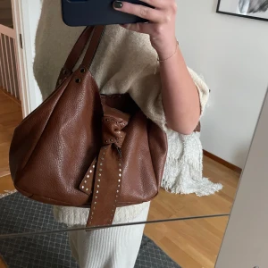 Brun axelväska med nitar - Säljer en snygg brun axelväska i skinn med coola nitar och en dekorativ knytning framtill. Perfekt för höst och vår! Väskan har en rymlig design och är i mycket bra skick. Passar både till vardags och festliga tillfällen.