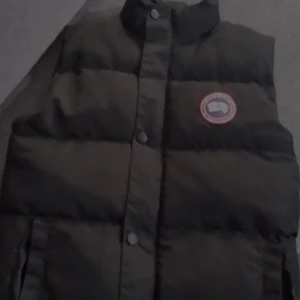 Svart dunväst från Canada Goose - Säljer en svart dunväst från Canada Goose i mycket bra skick. Västen har en broderad logga på bröstet och knappar framtill. Perfekt för höst och vinter, håller dig varm och stilren. Passar både till vardags och lite finare tillfällen.