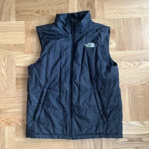 Svart dunväst från The North Face - Säljer en svart dunväst från The North Face i bra skick (ORIGINAL PRIS 1200kr) mitt pris under 50%. Västen har en dragkedja framtill och två praktiska fickor. Perfekt för vinter, höst och vår när du vill hålla dig varm utan att bära en jacka. Passar både för vardag och utomhusaktiviteter.