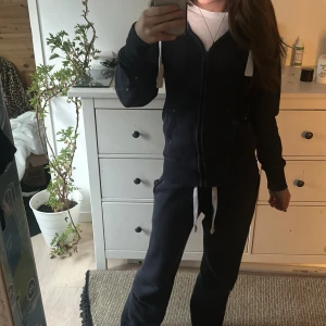 Svart hoodie med dragkedja - Säljer en mörkblå hoodie och byxor  med dragkedja på tröjan och vita snören. Perfekt för en avslappnad stil, den har långa ärmar och en bekväm passform. Passar bra till både vardag och träning. Hör av dig vid intresse!