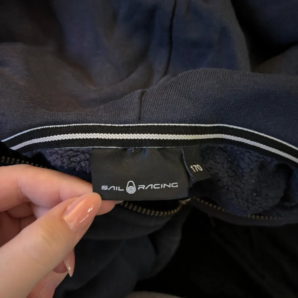 Säljer en mörkblå hoodie från Sail Racing i bra skick. Den har en dragkedja framtill och en broderad logga på bröstet och huvan. Perfekt för höst och vinter, och den är superbekväm att ha på sig. Passar både till vardags och lite mer avslappnade tillfällen.. Neuletakit & Villapaidat.