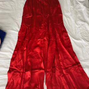 Röd långvida byxor i satin - Säljer en elegant röd långvid byxa i satin. Perfekt för festliga tillfällen med sin glänsande yta och flytande fall. Byxorna har en smickrande passform och är i mycket bra skick. Perfekt för att göra ett intryck på nästa fest eller bal!