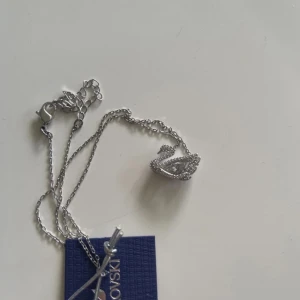 Silverfärgat halsband med svan från Swarovski - Säljer ett elegant silverfärgat halsband från Swarovski med en vacker svanberlock. Halsbandet är i mycket bra skick och kommer i originalförpackning. Perfekt för att lägga till en touch av glamour till vilken outfit som helst. Passar både till vardag och fest! Endast att förpackningen har gått sönder på sidan, som ni ser på bilden. 
