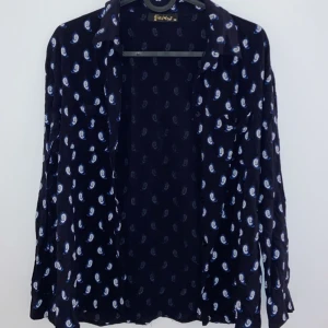 Svart paisleymönstrad skjorta från Savana - Snygg svart skjorta från Savana med ett stilrent paisleymönster i vitt. Perfekt för både vardag och fest! Skjortan har långa ärmar och en klassisk krage. Materialet känns lätt och bekvämt, vilket gör den idealisk för alla säsonger.