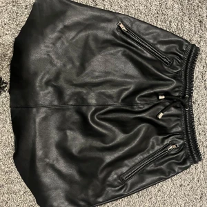 Svart kjol i skinnimitation från NA-KD - Snygg svart kjol i skinnimitation från NA-KD i storlek XS. Kjolen har en elastisk midja med dragsko och två coola dragkedjor framtill. Perfekt för en trendig look till både vardag och fest! 🖤