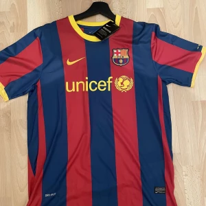 Fotbollströja Barcelona - Säljer en snygg fotbollströja från Nike i blått och rött med gula detaljer. Tröjan har korta ärmar och är i mycket bra skick. Den har ett randigt mönster och är perfekt för alla fotbollsfans. Passar både för träning och vardag. Dri-FIT material för bästa komfort. Perfekt för dig som älskar fotboll och vill visa ditt stöd! Skick 10/10. Aldrig använd. Prislappen sitter kvar. Tröjan är äkta. Storlek L. Bara att höra av sig vid minsta fundering!