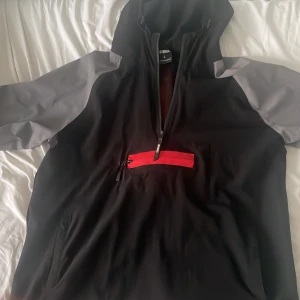 Svart och grå vindjacka från Hoodrich - Säljer en snygg svart och grå vindjacka från Hoodrich. Den har en cool röd detalj med dragkedja på framsidan och en stor röd logga på baksidan. Perfekt för höst och vår med sin lätta och bekväma passform. Jackan har en huva och långa ärmar, vilket gör den idealisk för blåsiga dagar. Storleken är L men passar också M bekvämt.