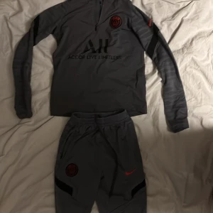 Psg Tracksuit - Säljer en grå Psg Tracksuit, långärmad tröja och matchande byxor. Tröjan har en halv dragkedja och ett tryckt emblem på bröstet. Perfekt för fotbollsträning eller annan sportaktivitet. Setet är i bra skick och redo för nya äventyr!