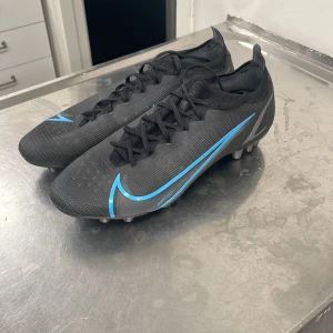 Svarta Nike mercurial fotbollsskor med blå detaljer - Säljer ett par svarta Nike fotbollsskor i syntetmaterial med snygga blå detaljer. Skorna har snörning och är i mycket bra skick. Perfekta för fotbollsträning eller match. De har en tight passform och är designade för att ge bra grepp på planen.