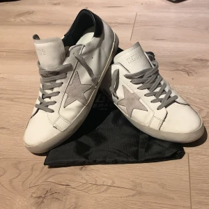 Golden Goose - Säljer ett par snygga vita sneakers med en grå stjärna på sidan. De har grå snörning och en svart detalj vid hälen. Perfekta för både vardag och fest! Skorna är i bra skick och kommer med originalpåse och låda.