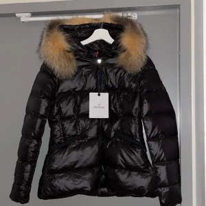 Svart dunjacka från Moncler - Säljer en snygg svart dunjacka från Moncler i mycket bra skick. Använd fåtal gånger den har en  fluffig äkta päls på luvan som går att ta bort och dragkedja framtill med ett bälte som går att ta bort. Jackans storlek är en M men är väldigt liten i storleken och passar en S.