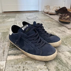 Blå sneakers från Golden Goose - Säljer ett par snygga blå sneakers från Golden Goose i bra skick. De har en cool stjärndetalj på sidan och vit häl med märkesnamnet. Skorna har en vintage-look med lite slitage på sulan, vilket ger dem en unik stil. Perfekta för både vardag och fest!