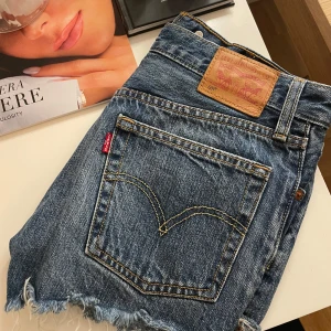 Jeansshorts från Levi's - Säljer ett par klassiska Levi's 501 jeansshorts i blå denim. De har en snygg fransig kant och knappar framtill. Perfekta för sommaren och ger en avslappnad look.