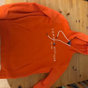 Orange hoodie från Tommy Hilfiger - Säljer en snygg orange hoodie från Tommy Hilfiger med vit text och logga på framsidan. Den har en klassisk känguruficka och justerbar huva med vita snören. Perfekt för en avslappnad stil och passar bra till höst och vår.