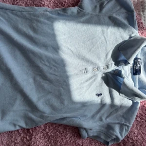 Ljusblå pikétröja från Ralph Lauren - Säljer en snygg ljusblå pikétröja från Ralph Lauren. Den har en klassisk krage och tre knappar framtill. Perfekt för en avslappnad men stilren look. Tröjan är i bra skick och passar både till vardags och lite finare tillfällen.
