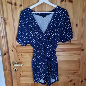 Blå prickig omlottklänning - Säljer en superfin blå omlottjumpsuit med vita prickar. Den har en v-ringad design och korta ärmar, perfekt för sommardagar. Klänningen har ett knytband i midjan för en snygg passform. Materialet känns lätt och luftigt, vilket gör den idealisk för både vardag och fest!