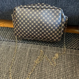 Mönstrad axelväska med kedja - Säljer en snygg axelväska med ett coolt mönster i brunt och guld. Väskan har en kedja som axelrem och en praktisk dragkedja upptill. Serr liten ut men får plats med extremt mycket i!