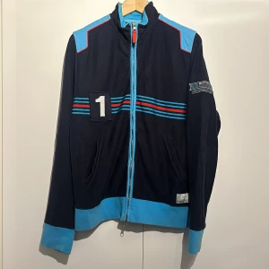 Blå jacka från Porsche Design - Säljer en snygg blå Cardigan från Porsche Design med coola Martini Racing detaljer. Tröjan har en dragkedja framtill och är långärmad. Den har ett broderat nummer '1' på framsidan och texten 'MARTINI RACING' på baksidan. Perfekt för höst och vår!