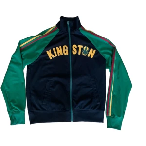 Svart och grön jacka från Lucky Brand - Säljer en cool svart och grön jacka från Lucky Brand med texten 'KINGSTON' i gult på framsidan. Jackan har långa ärmar med ränder i rött och gult längs ärmarna. Den är gjord av en blandning av bomull och polyester och har en dragkedja framtill. Perfekt för en avslappnad stil!