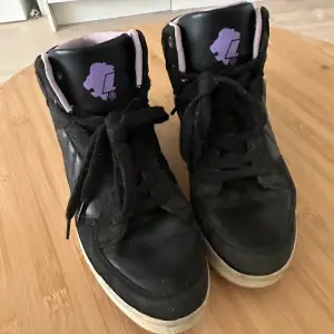 Säljer ett par svarta sneakers med lila detaljer. Skorna har en hög modell med snörning och en vit sula. De är i välanvänt skick men fortfarande snygga och bekväma. Perfekta för vardagsbruk eller en avslappnad stil.