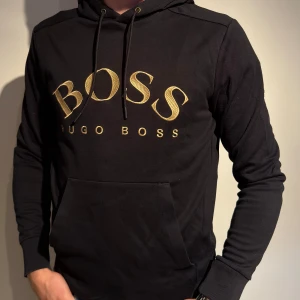 Svart hoodie från Hugo Boss - Säljer en snygg svart hoodie från Hugo Boss med guldtext på bröstet. Den har en klassisk känguruficka och justerbar huva med snören. Perfekt för en stilren och bekväm look!