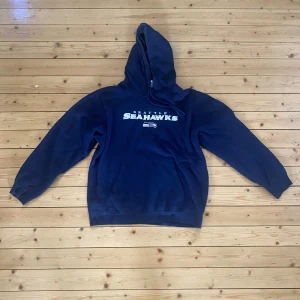 Blå Seattle Seahawks hoodie - Säljer en snygg blå hoodie med Seattle Seahawks-logga på bröstet. Perfekt för kalla dagar med sin mysiga huva och långa ärmar. Den är i bra skick och passar perfekt för en avslappnad stil.