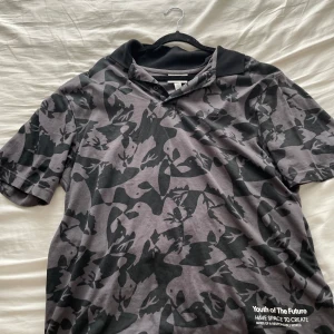 Grå och svart camouflagemönstrad t-shirt - Säljer en grå och svart camouflagemönstrad t-shirt från Youth of The Future. Den har korta ärmar och en klassisk krage. Perfekt för en avslappnad stil eller för att sticka ut lite extra. Texten 'Have Space to Create' ger en cool touch. Passar bra till både jeans och shorts!