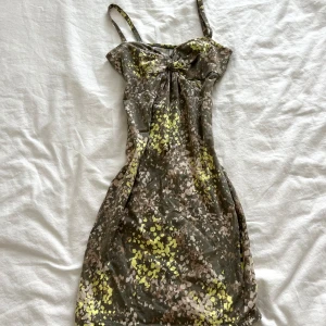 Camouflage klänning med rosett - Säljer en supercool klänning med camouflage-mönster i gröna och gula toner. Den har smala axelband och en söt rosett framtill. Perfekt för sommarens fester eller en dag på stan. Klänningen är i ett mjukt och bekvämt material.