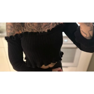 Svart ribbad offshoulder topp - Svart ribbad offshoulder topp. Den är croppad och har långa ärmar med volangdetaljer vid ärmslut och nederkant. Storlek S!