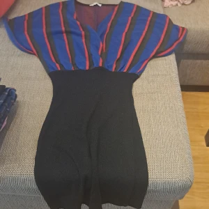 Randig klänning i blått och rött - Säljer en snygg klänning med randigt mönster i blått, rött och svart. Klänningen har en v-ringad överdel med korta ärmar och en tight passform i midjan som ger en fin siluett. Perfekt för både vardag och fest! oanvänd. 100% nytt. 