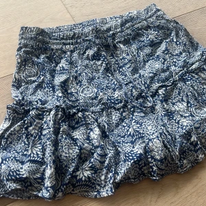 Blå och vit blommig kjol från Zara - Säljer en superfin blå och vit blommig kjol från Zara. Den har en volangdetalj som ger den en söt och lekfull look. Perfekt för sommardagar eller en avslappnad vårutflykt. Kjolen är i storlek S och har en bekväm passform. Har inbyggda shorts men syns inte ❤️